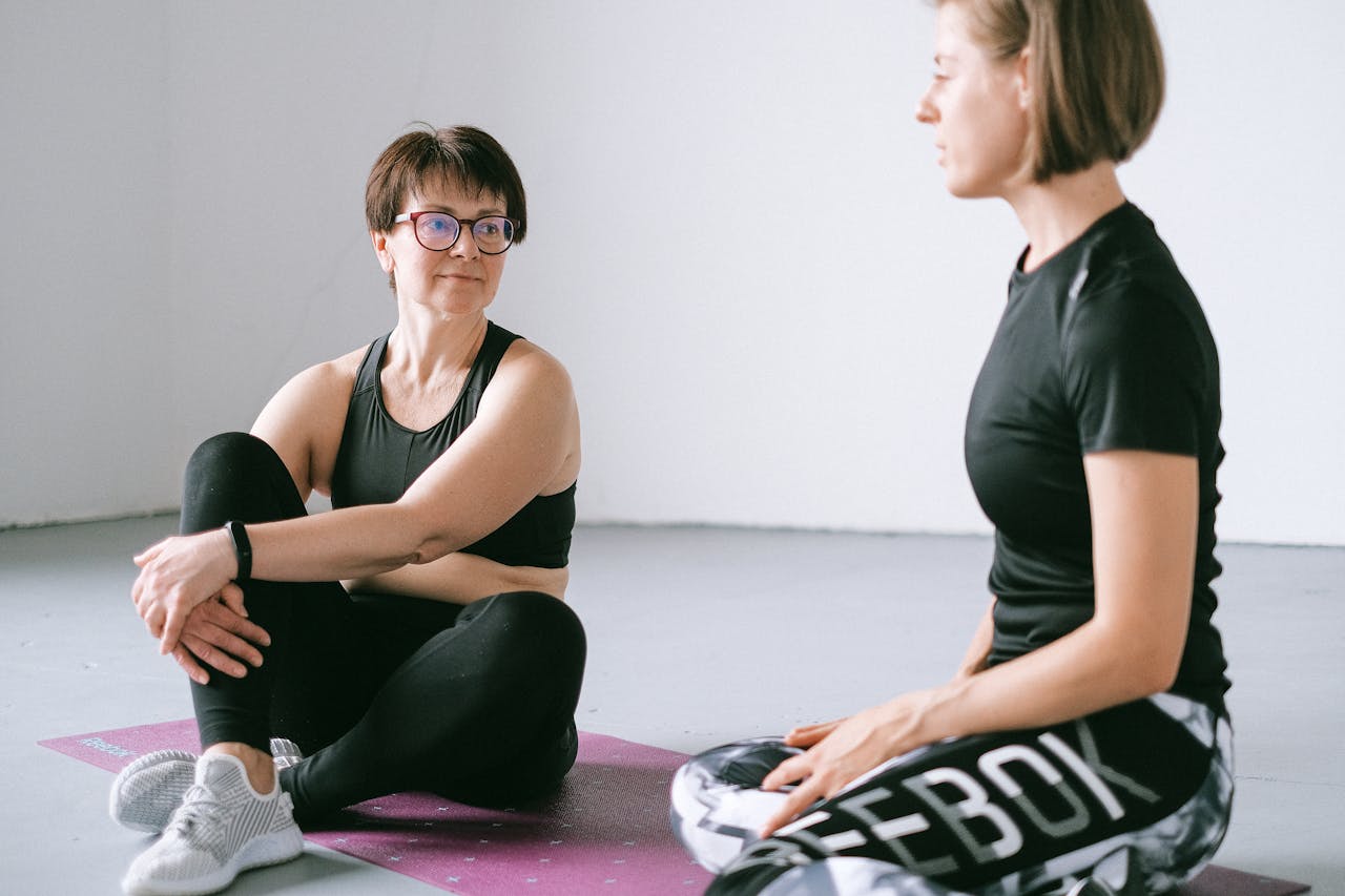 Two women in activewear discuss fitness on a yoga mat indoors.