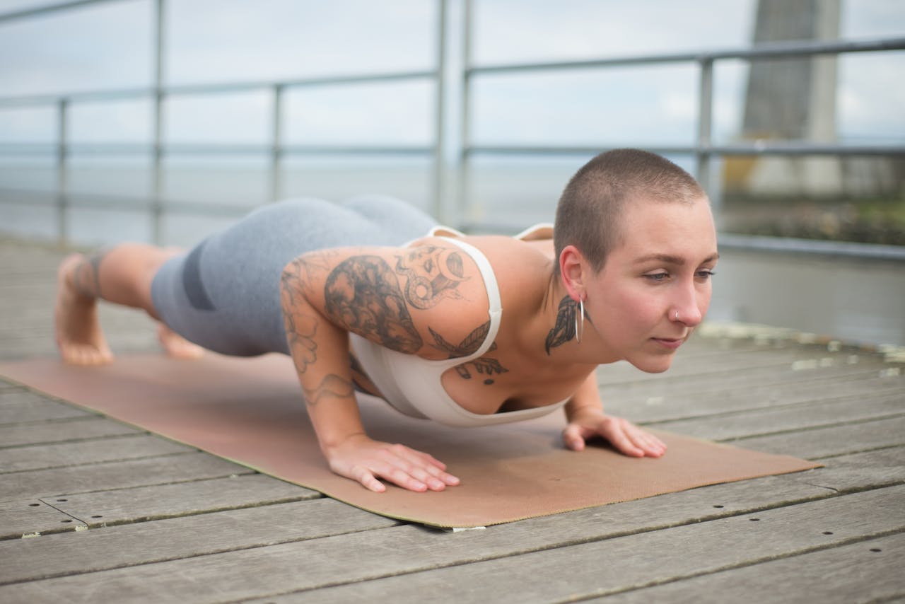 Bald woman in sportswear performing yoga posture on a wooden deck by the sea, showcasing tattoos.